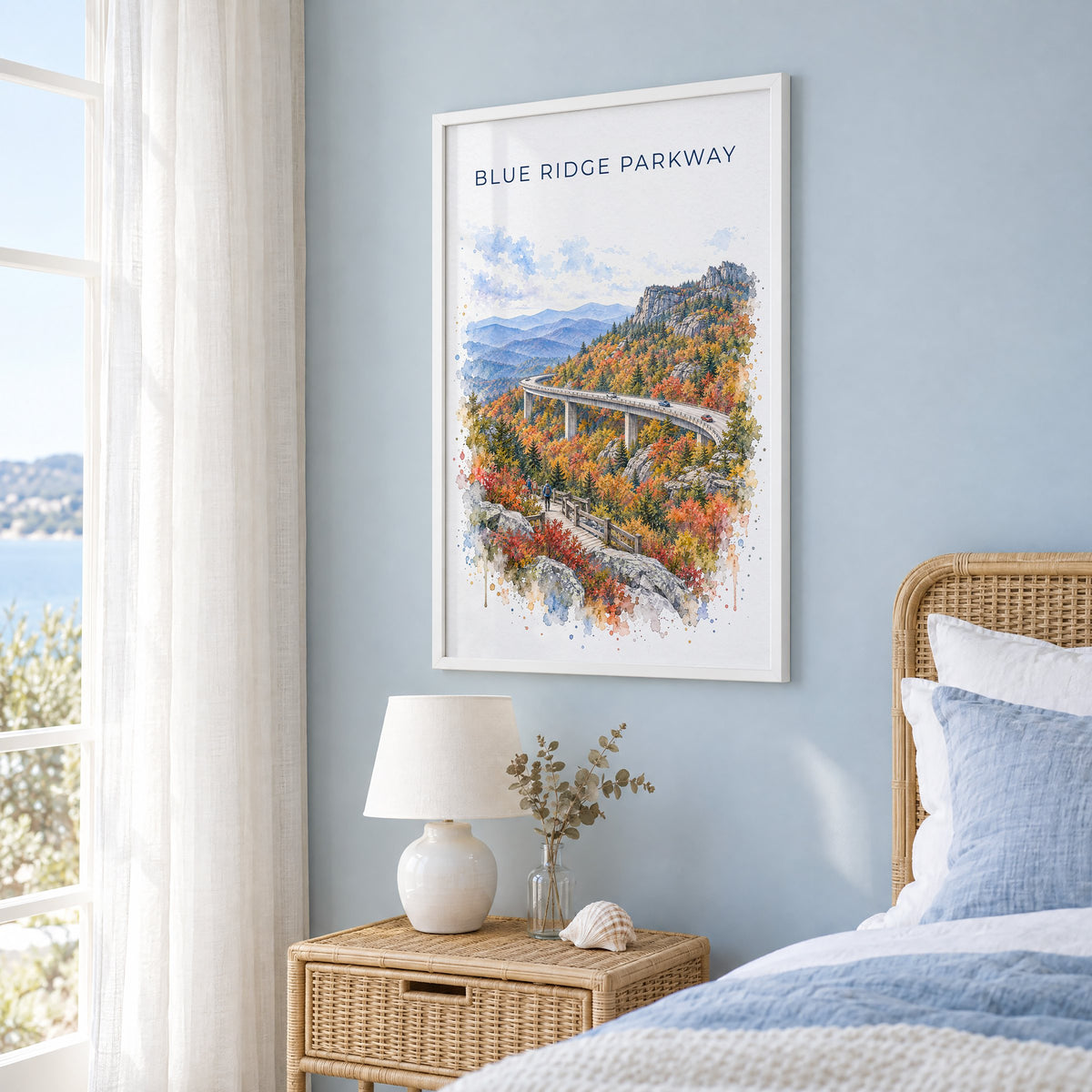 Blue Ridge Parkway watercolor print in a white frame on a pale blue wall beside a rattan bed in a coastal bedroom