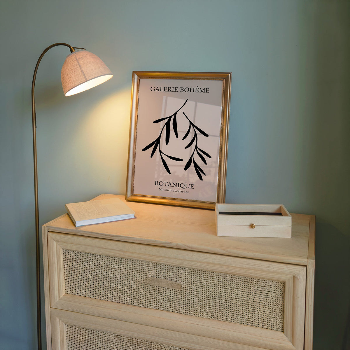 Two black hanging branches on blush botanical print shown in a gold frame on a cane dresser lit by a floor lamp