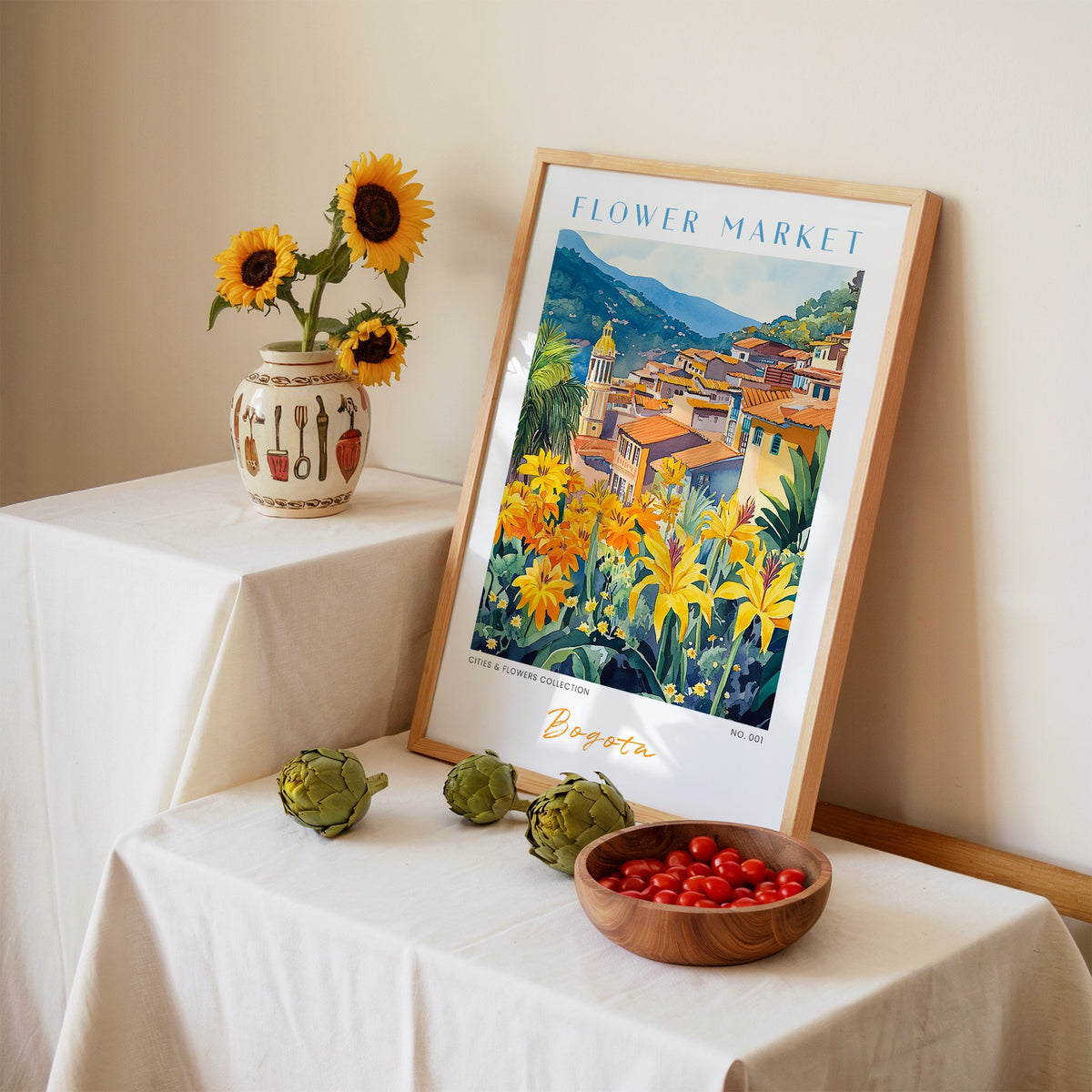 Bogota flower market print in an oak frame leaning on a linen-draped table beside a vase of sunflowers