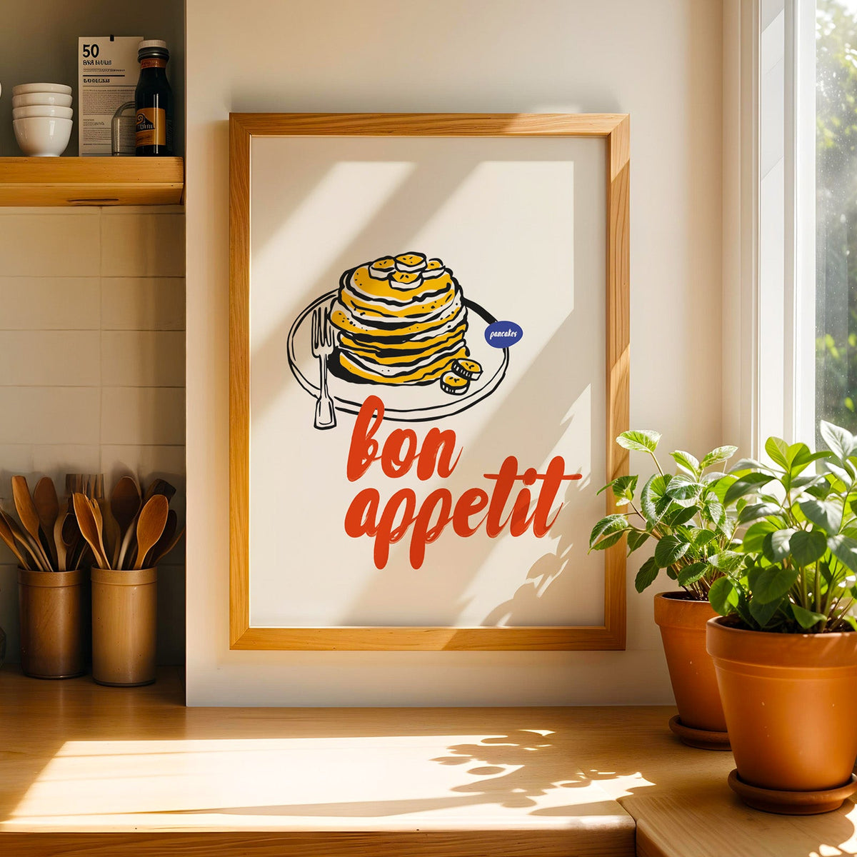 Bon Appetit pancakes print in a light pine frame beside a jar of wooden spoons