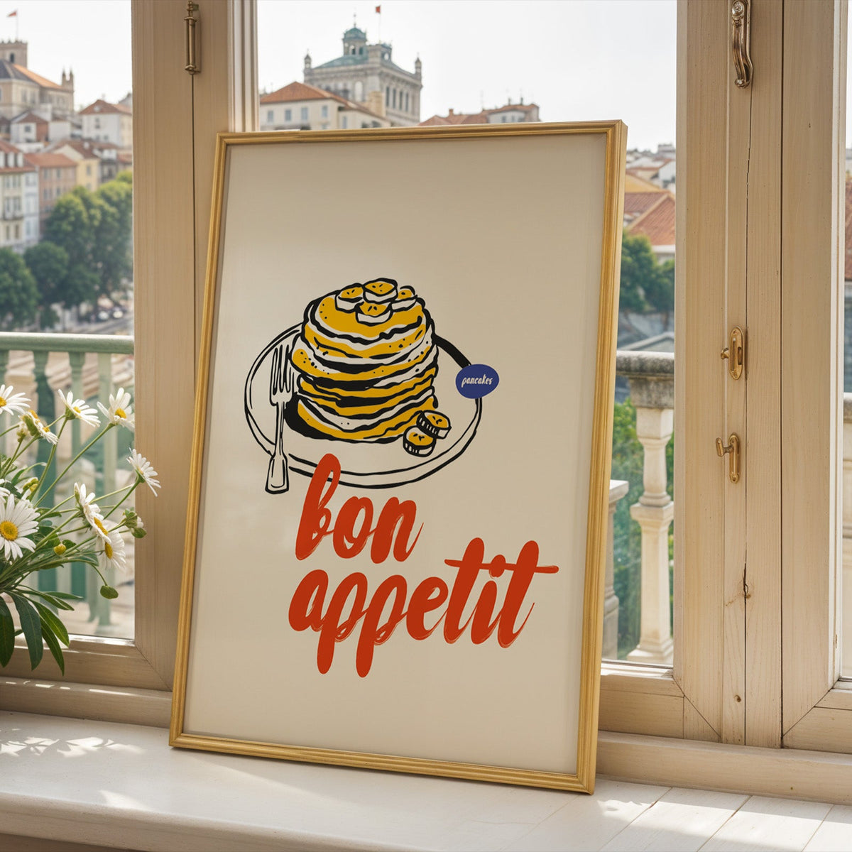 Bon Appetit pancakes print in a gold frame on a window sill above a hillside town