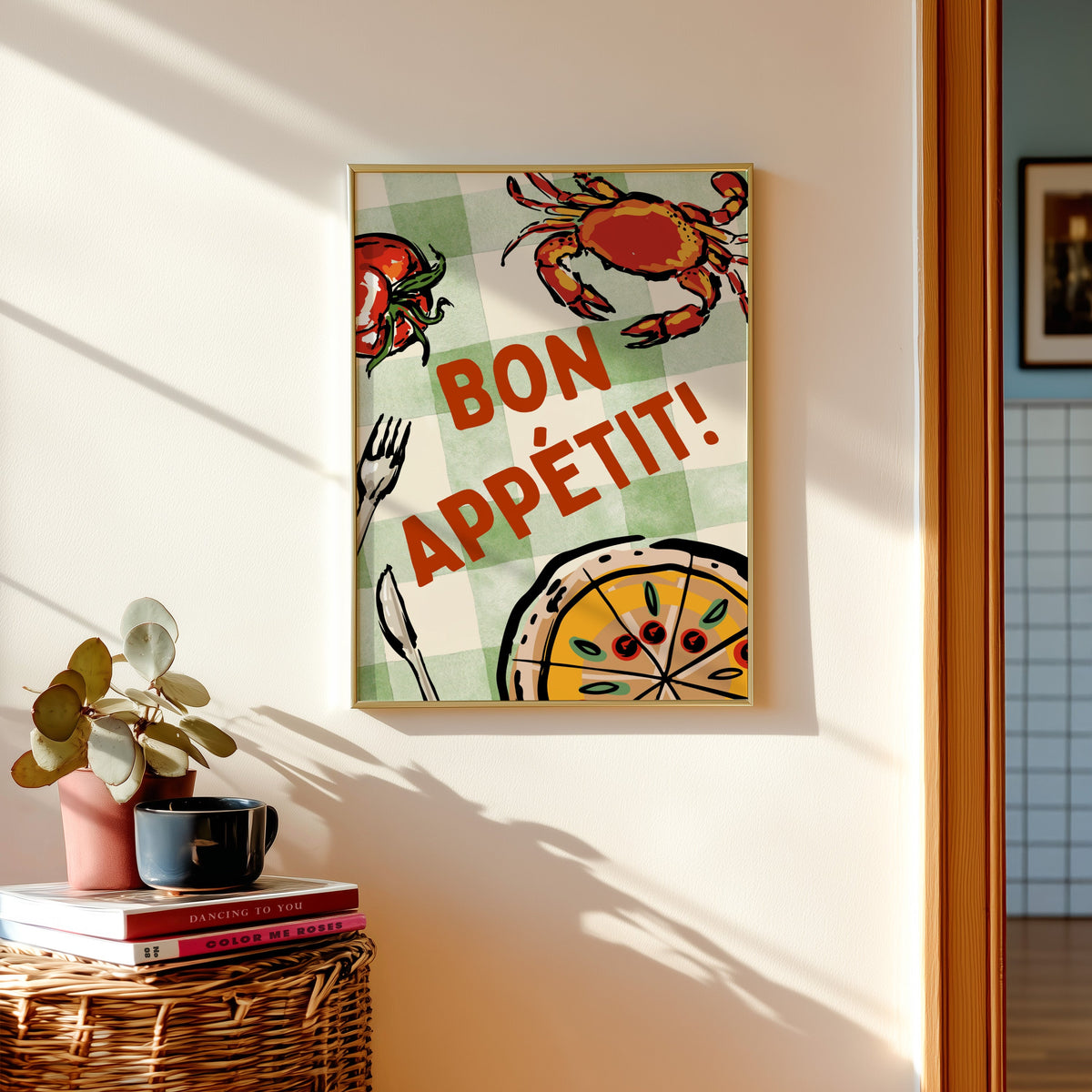 Bon Appetit crab print shown in a gold frame above a basket of books and a pilea plant