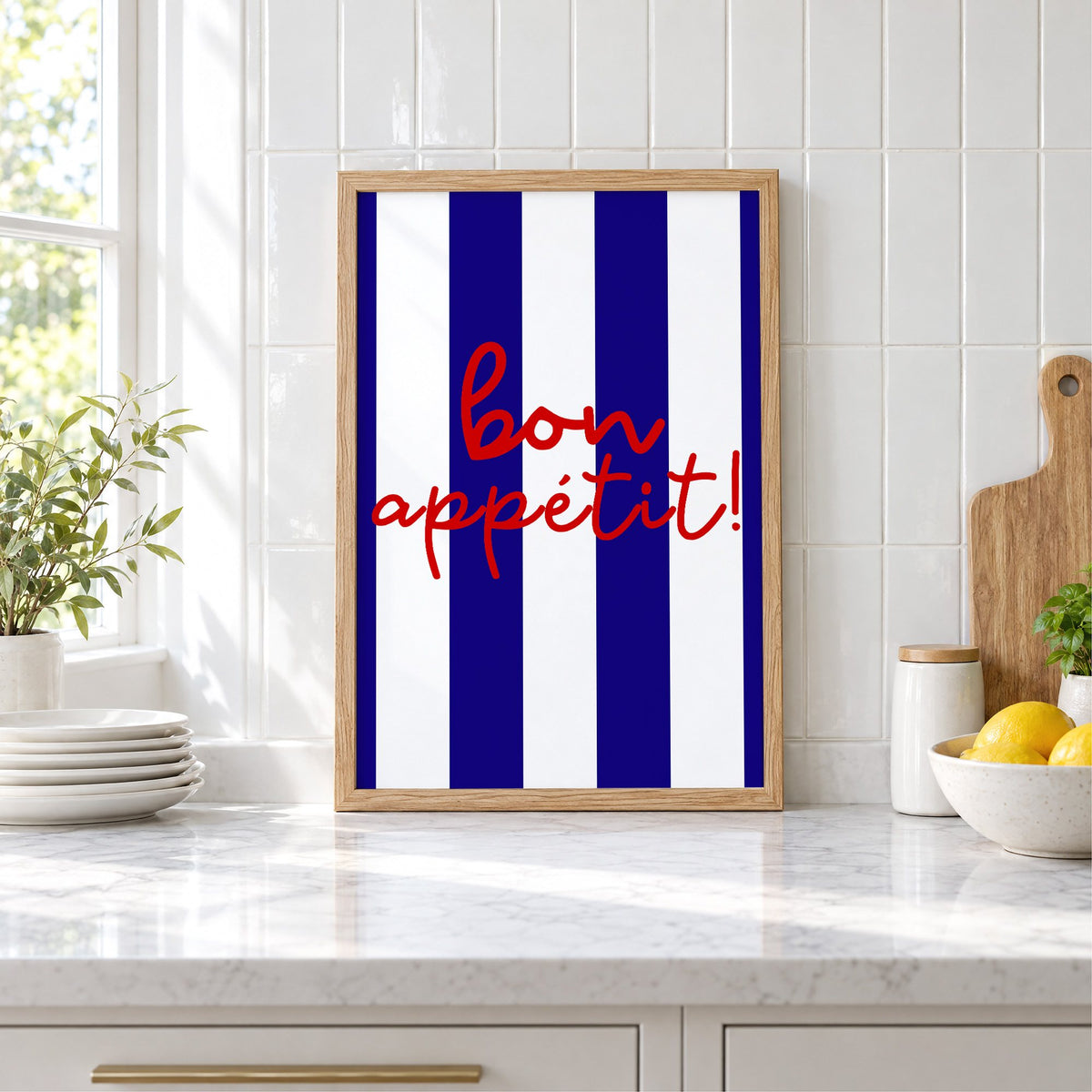 Bon appétit navy stripe print in an oak frame leaning on a marble counter by white tiles and lemons