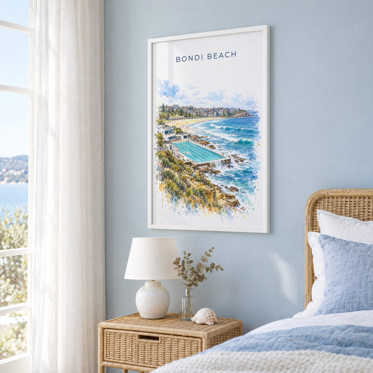 Bondi Beach watercolor print in a white frame on a pale blue wall beside a rattan bed in a coastal bedroom