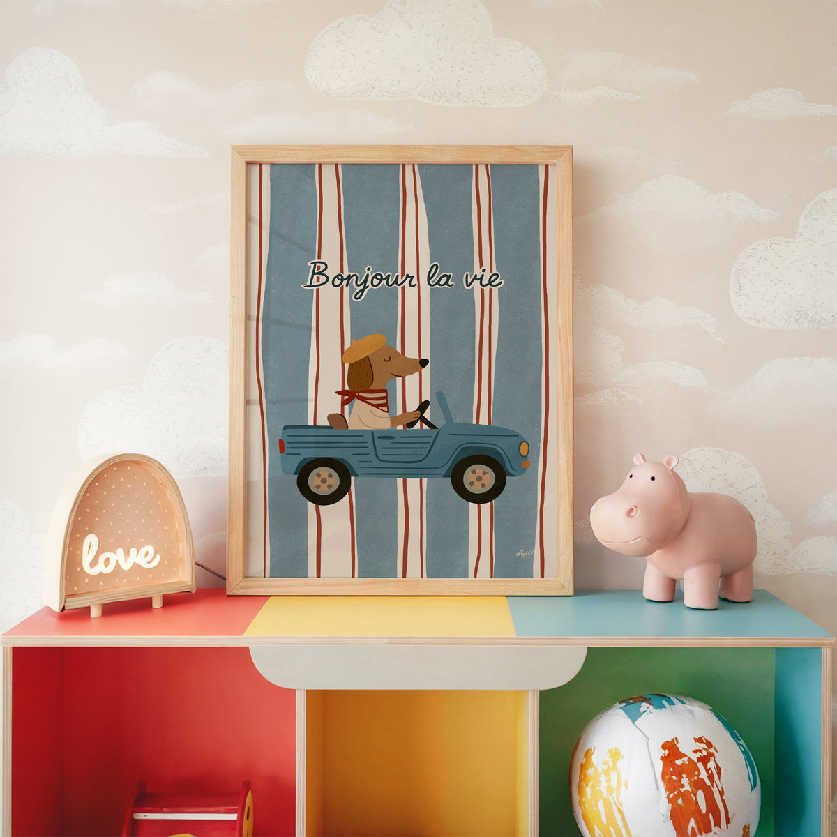 Dog driving car with french script nursery print in an oak frame on a rainbow toy shelf against cloud wallpaper