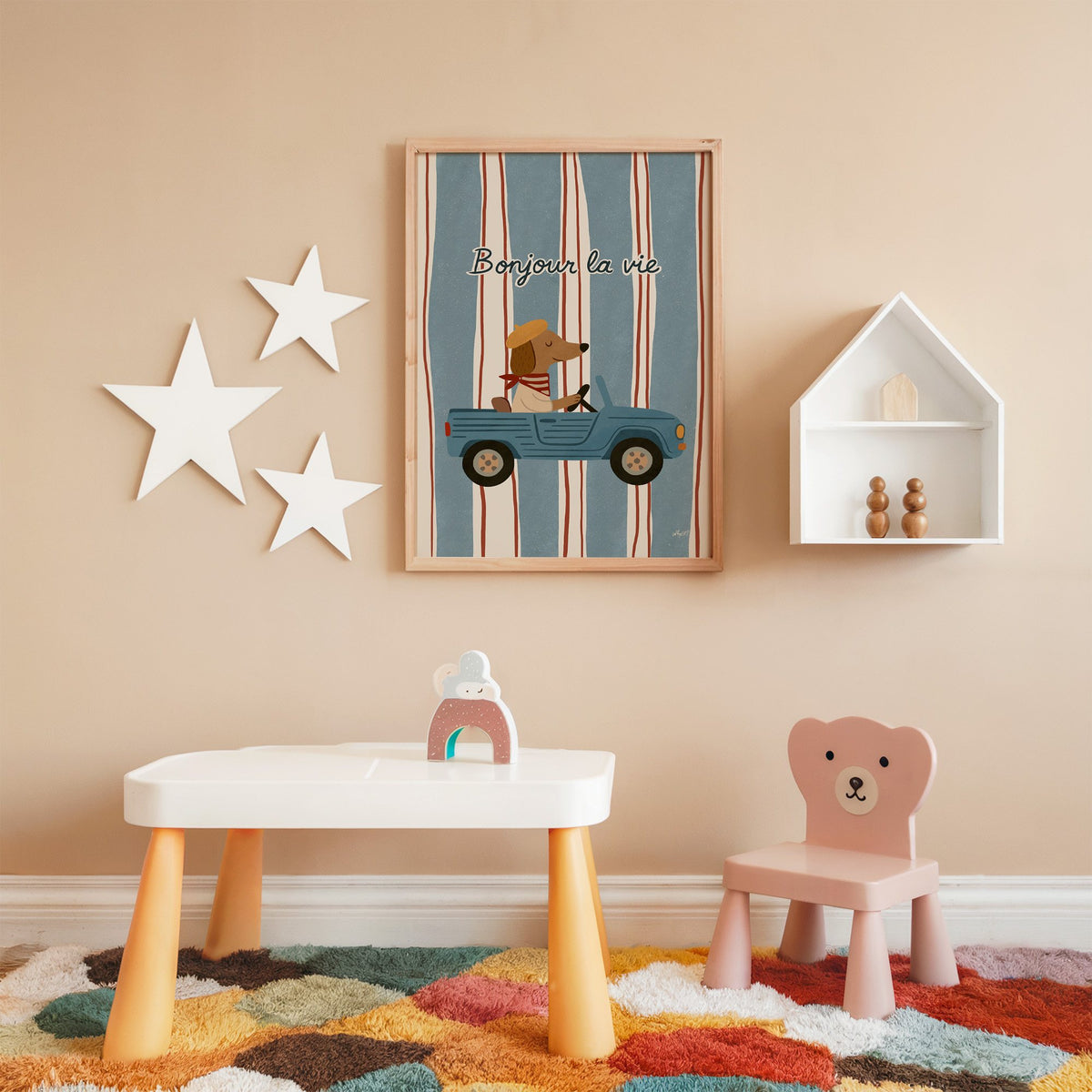 Dog driving car with french script nursery print in an oak frame on a peach wall above a kids table with wall stars