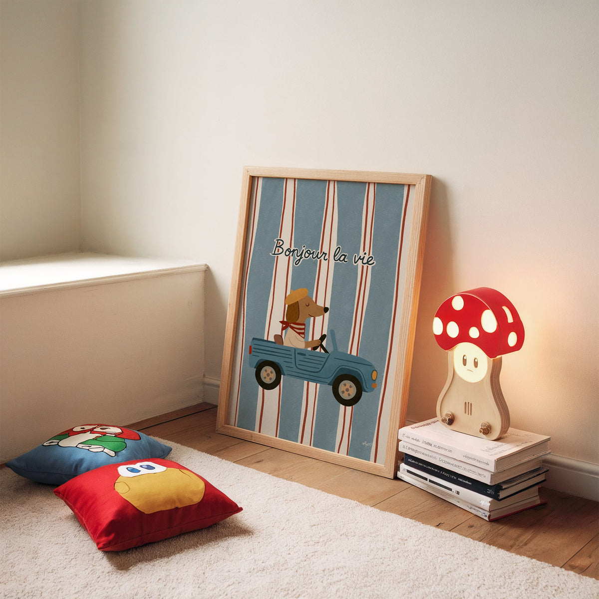 Dog driving car with french script nursery print in an oak frame leaning on a rug beside a mushroom lamp and floor cushions