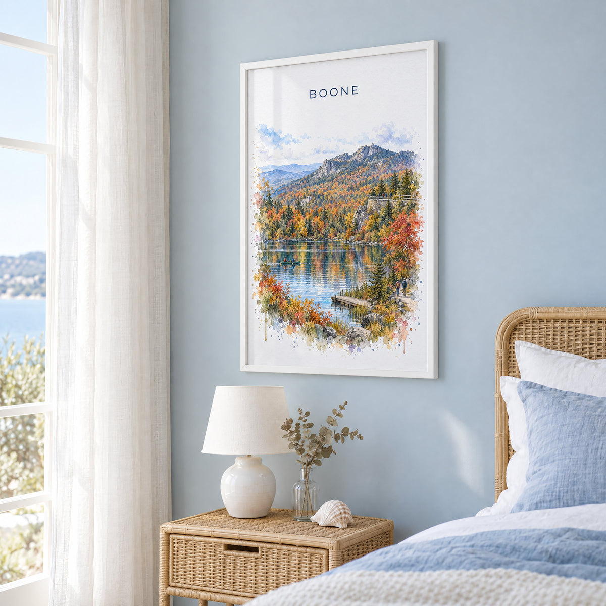 Boone, North Carolina watercolor print in a white frame on a pale blue wall beside a rattan bed in a coastal bedroom