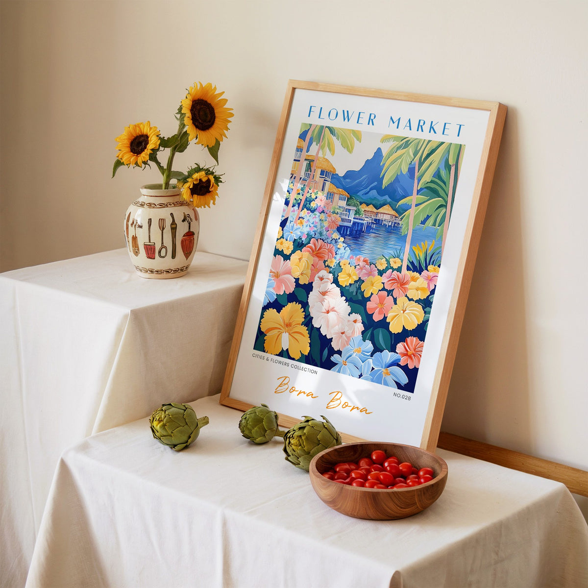 Bora Bora flower market print in an oak frame leaning on a linen-draped table beside a vase of sunflowers