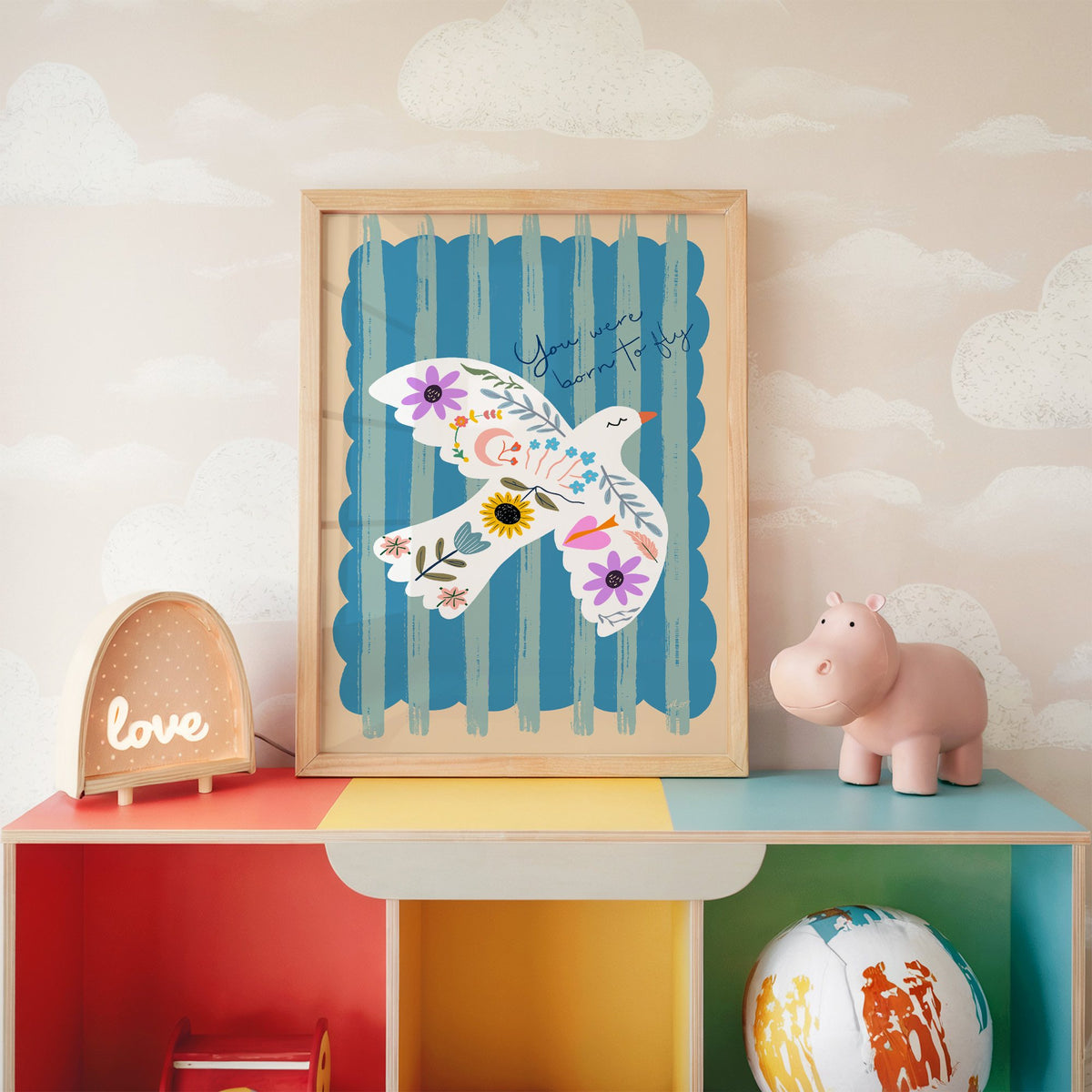 White dove covered in flowers nursery print in an oak frame on a rainbow toy shelf against cloud wallpaper