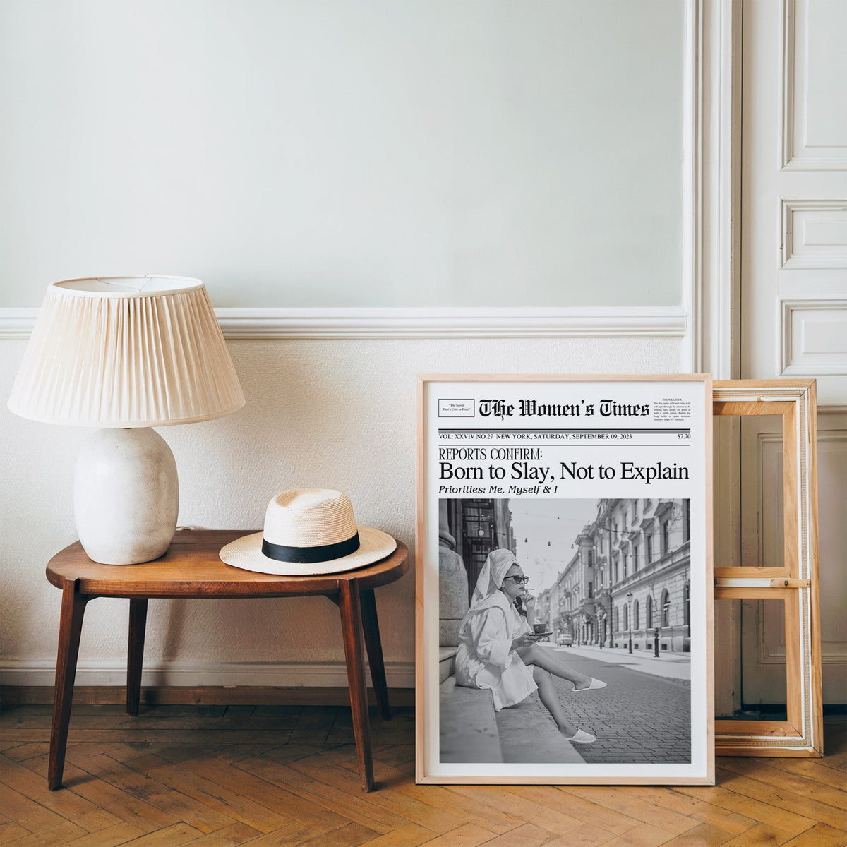 Mock front page with woman in robe on a cobbled street print in a pine frame on a wood floor beside a lamp and straw hat