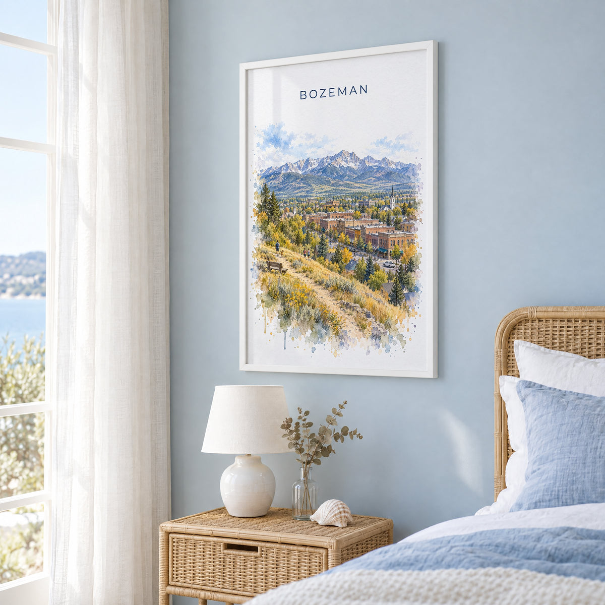 Bozeman watercolor print in a white frame on a pale blue wall beside a rattan bed in a coastal bedroom