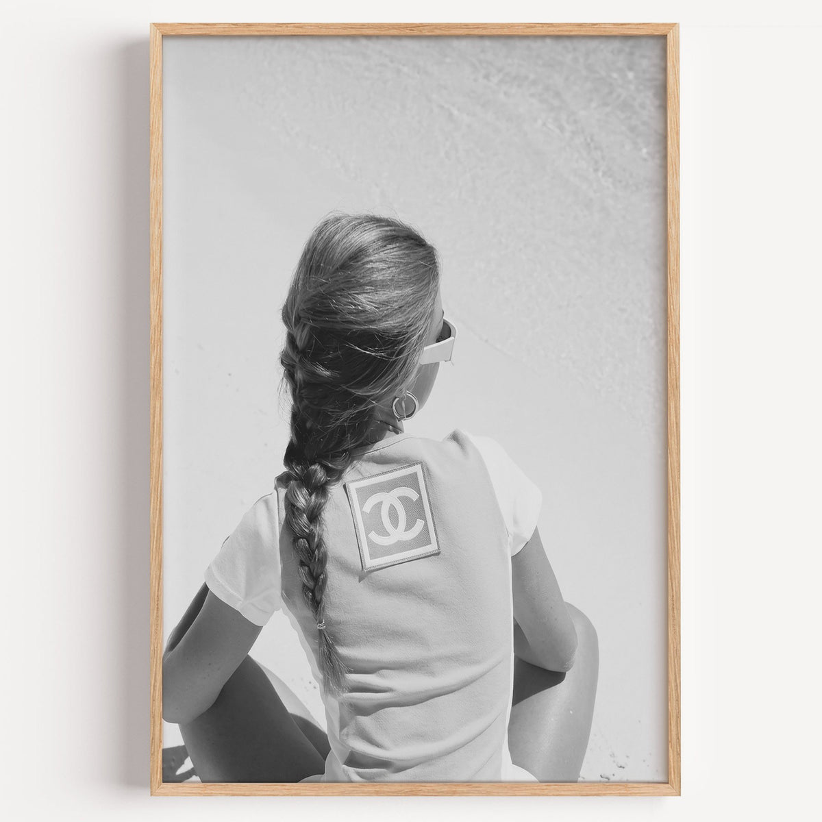 Woman with long braid seated on beach from behind print in a light oak frame on a plain white wall