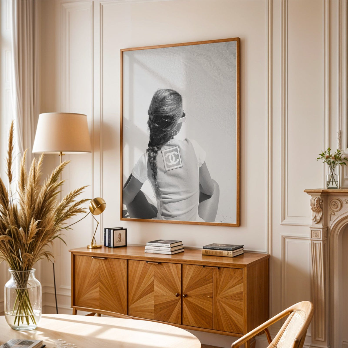 Woman with long braid seated on beach from behind print in an oak frame above a sunburst wood sideboard with pampas grass