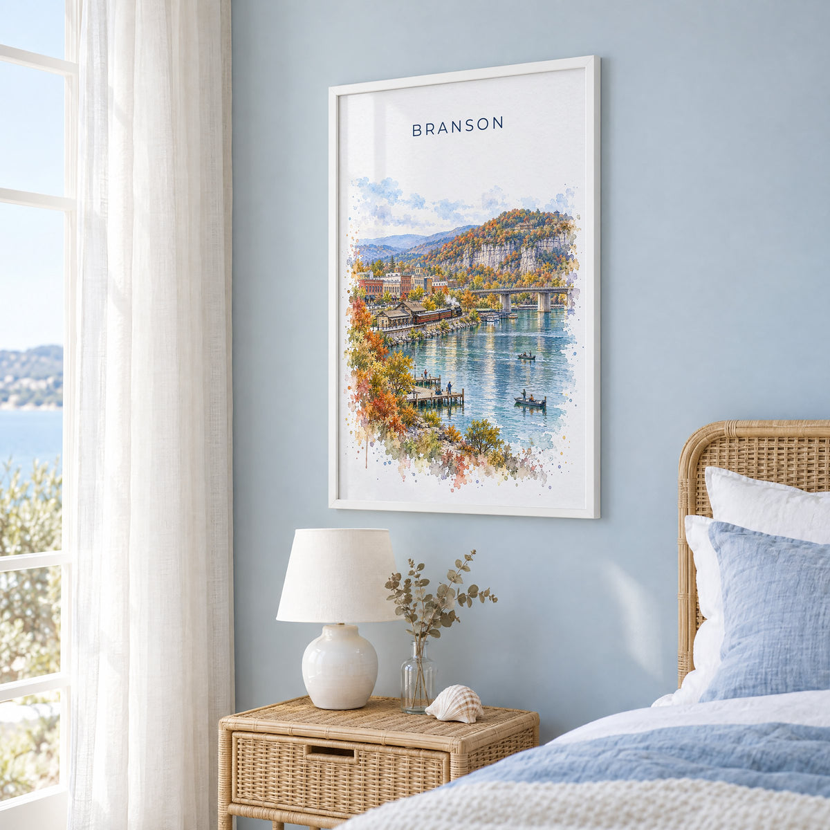 Branson watercolor print in a white frame on a pale blue wall beside a rattan bed in a coastal bedroom