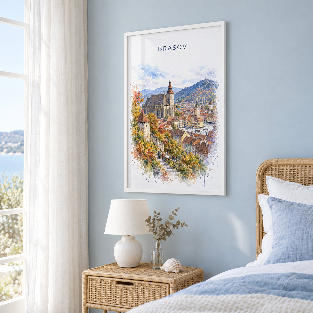 Brasov watercolor print in a white frame on a pale blue wall beside a rattan bed in a coastal bedroom