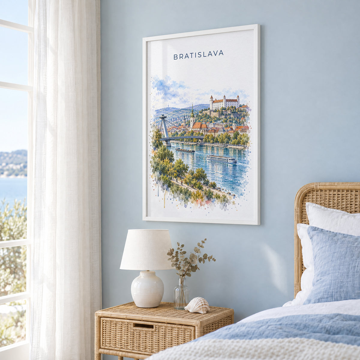 Bratislava watercolor print in a white frame on a pale blue wall beside a rattan bed in a coastal bedroom