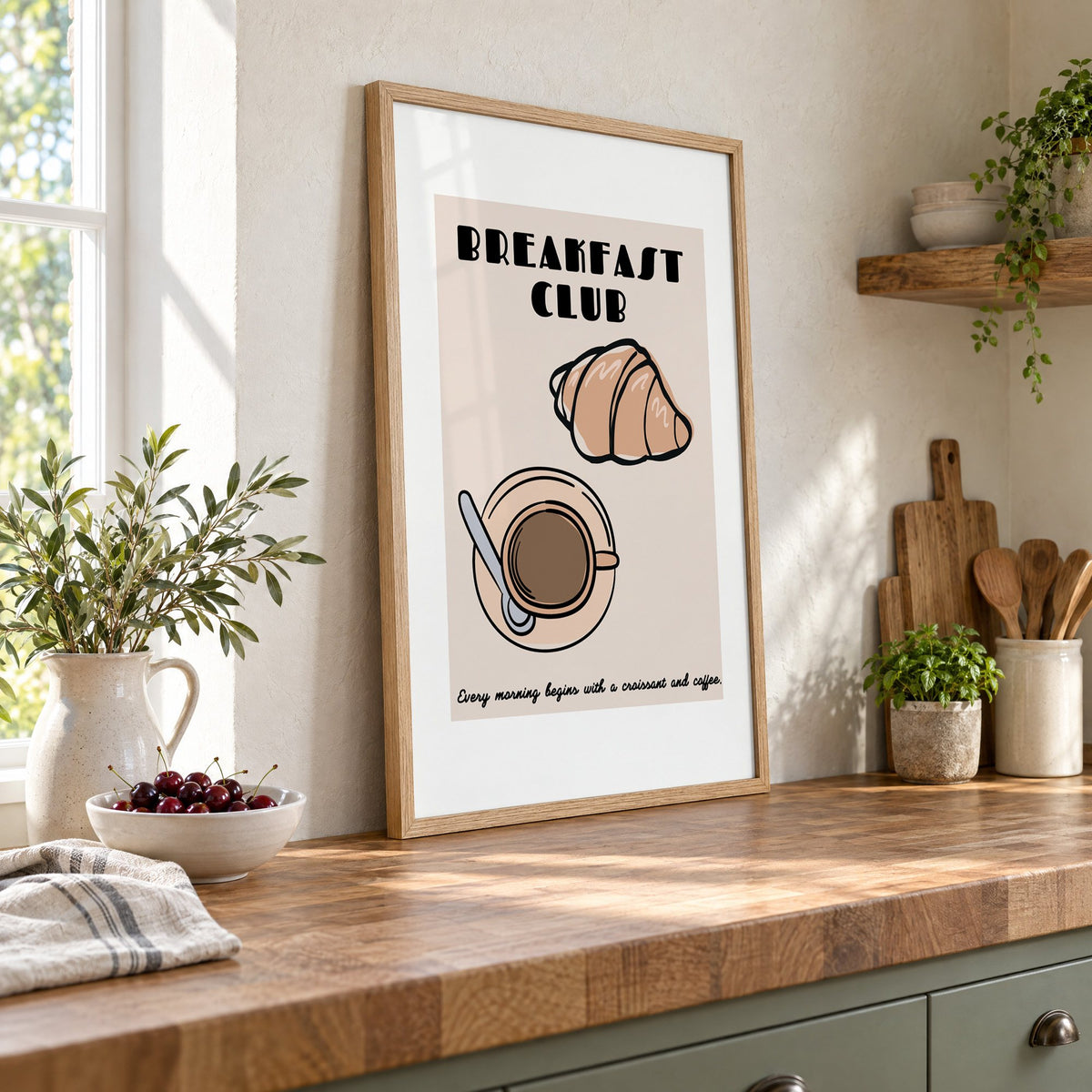 Breakfast Club croissant and coffee print in an oak frame leaning on a butcher block island beside a bowl of cherries