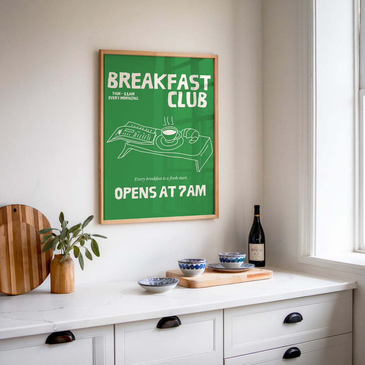 Breakfast Club green tray print in an oak frame above a white kitchen sideboard with a wine bottle and wooden board