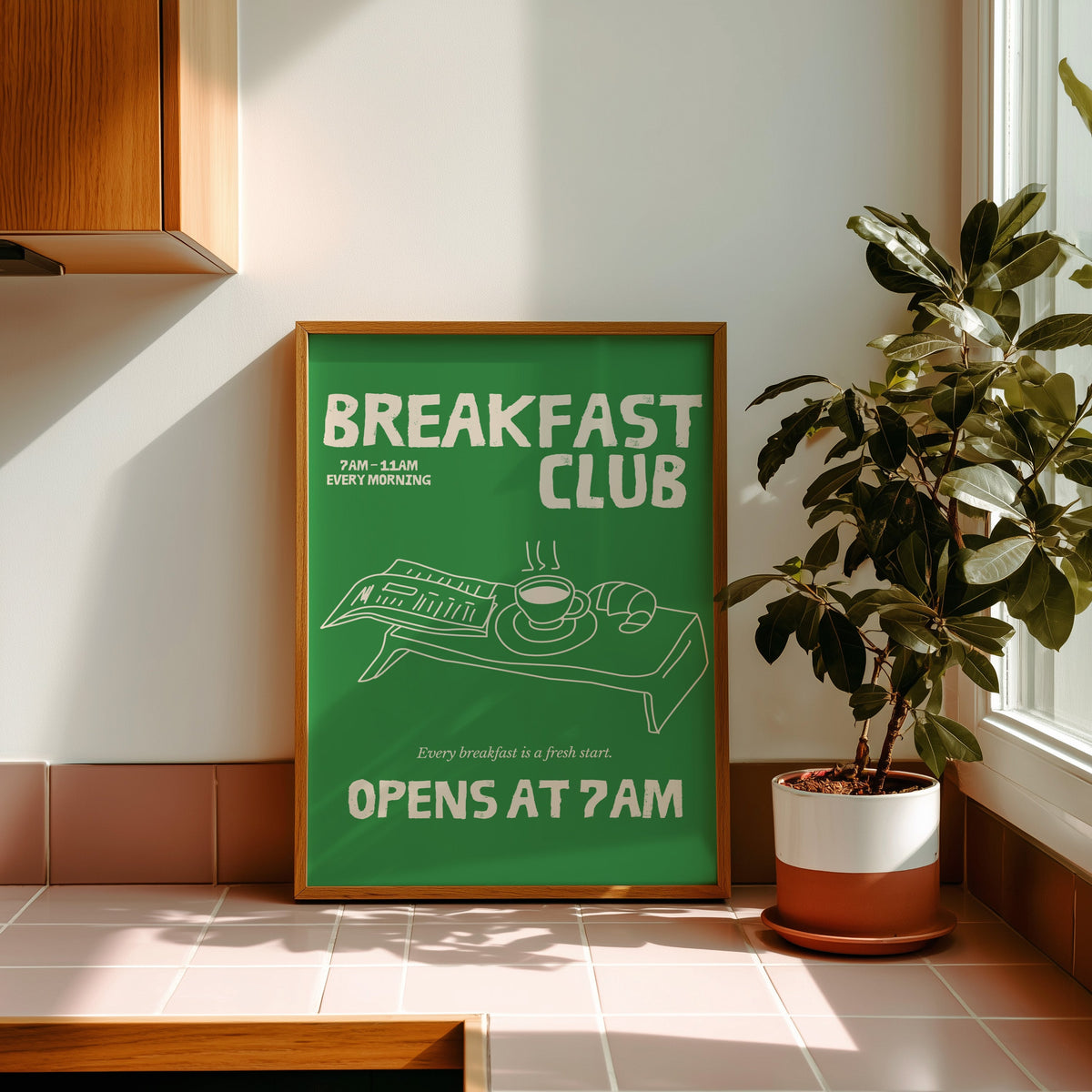 Breakfast Club green tray print in a golden oak frame leaning on a pink tiled ledge in strong afternoon sun