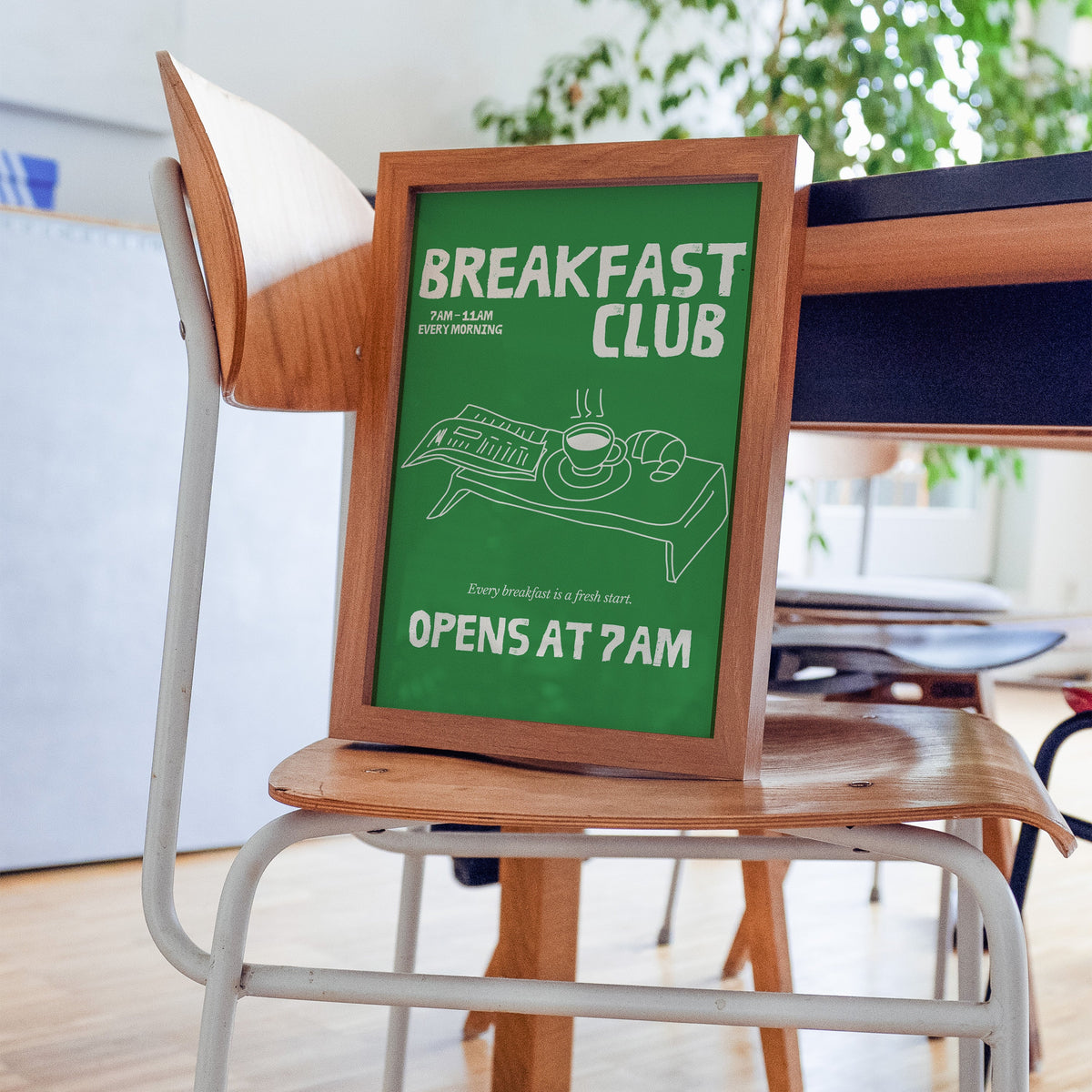 Breakfast Club green tray print in a walnut frame resting on a plywood studio chair beside a dark desk