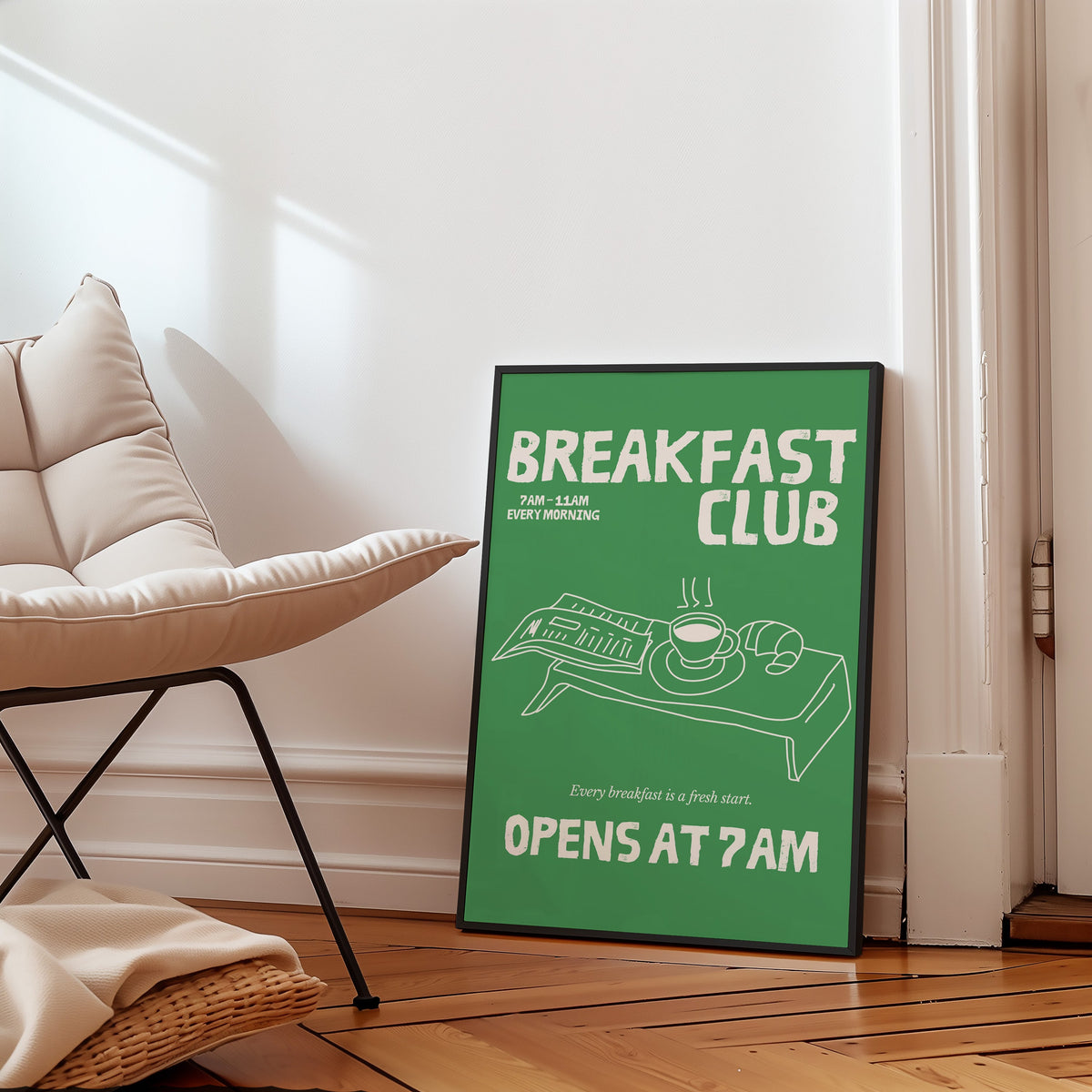 Breakfast Club green tray print in a slim black frame leaning on a herringbone floor beside a cream armchair