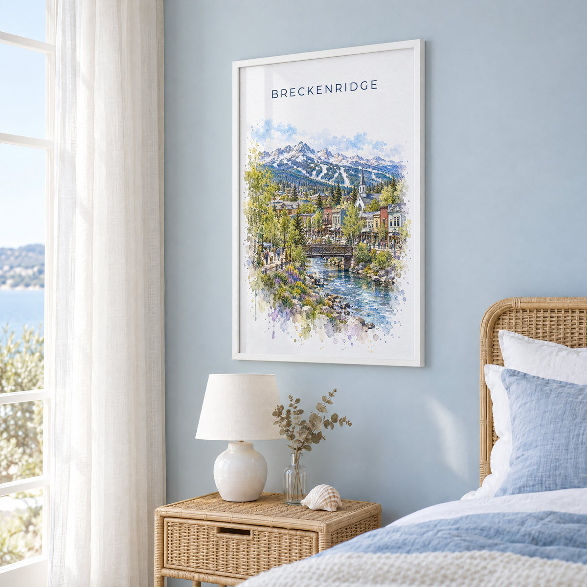 Breckenridge watercolor print in a white frame on a pale blue wall beside a rattan bed in a coastal bedroom