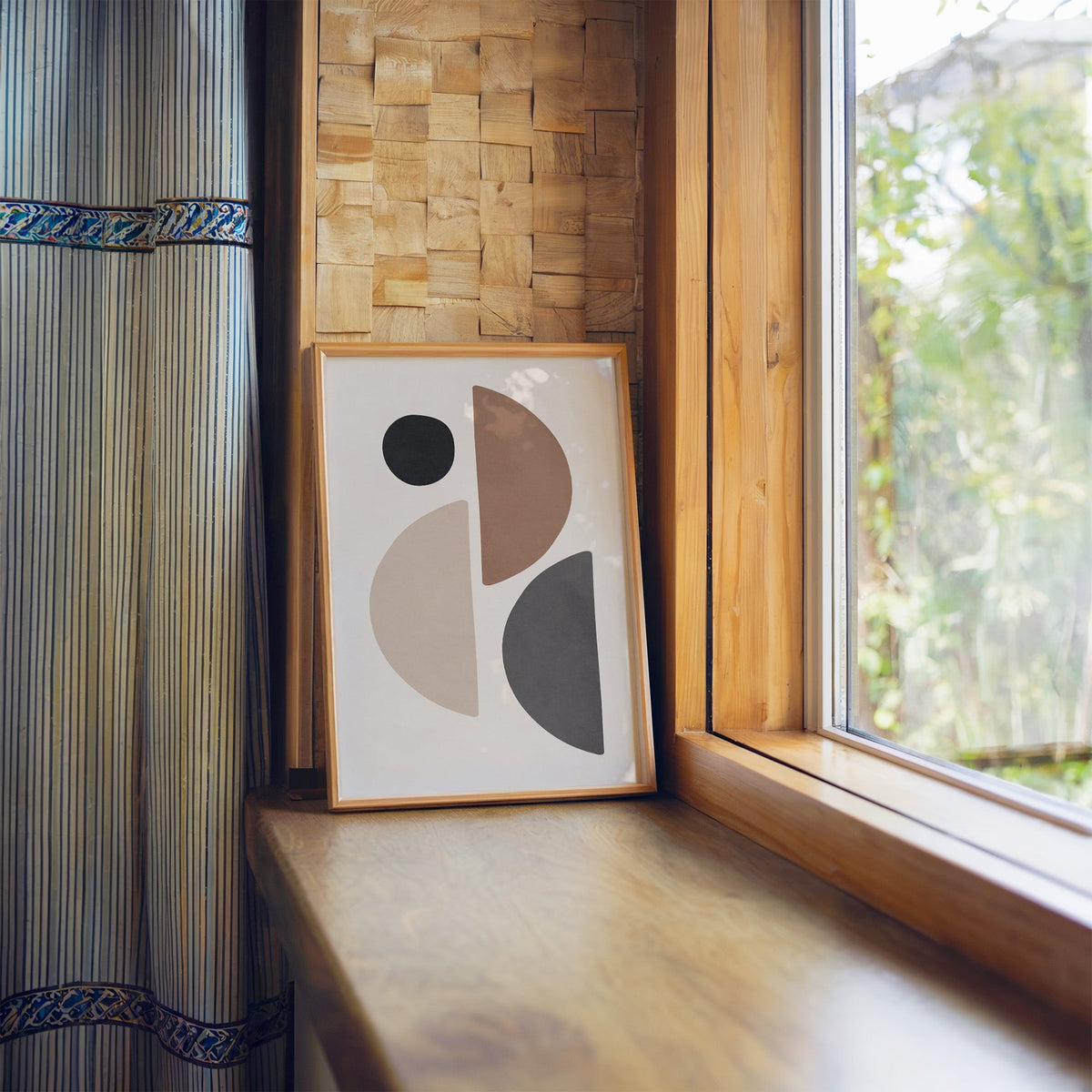Brown, grey and black split circle shapes print in a light wood frame leaning on a wooden window sill by a garden view
