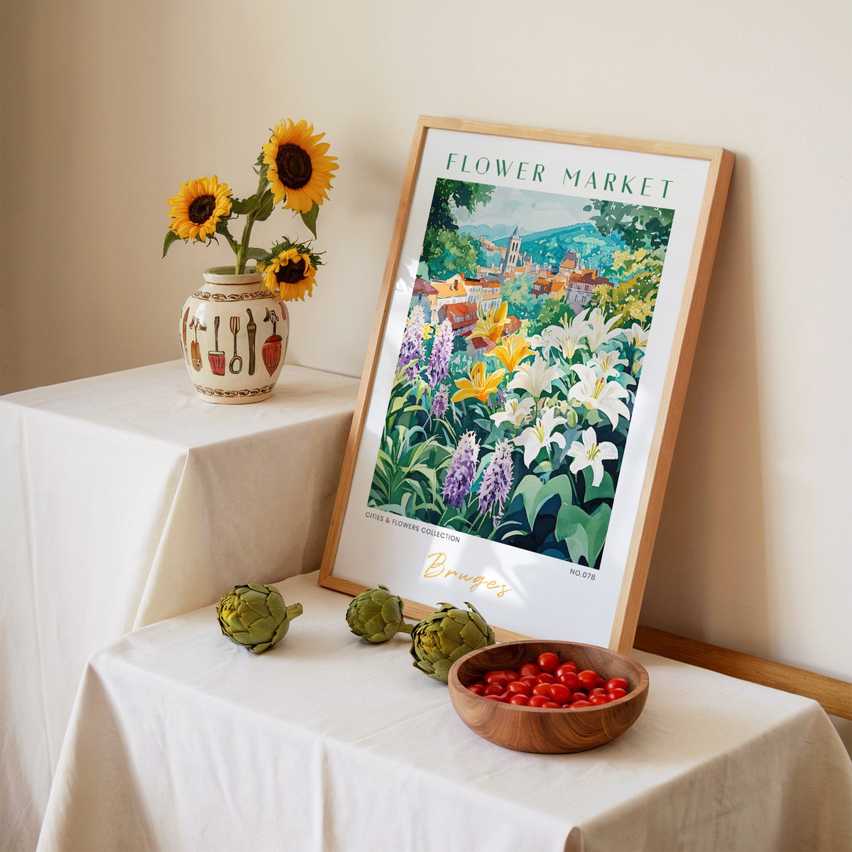 Bruges flower market print in an oak frame leaning on a linen-draped table beside a vase of sunflowers