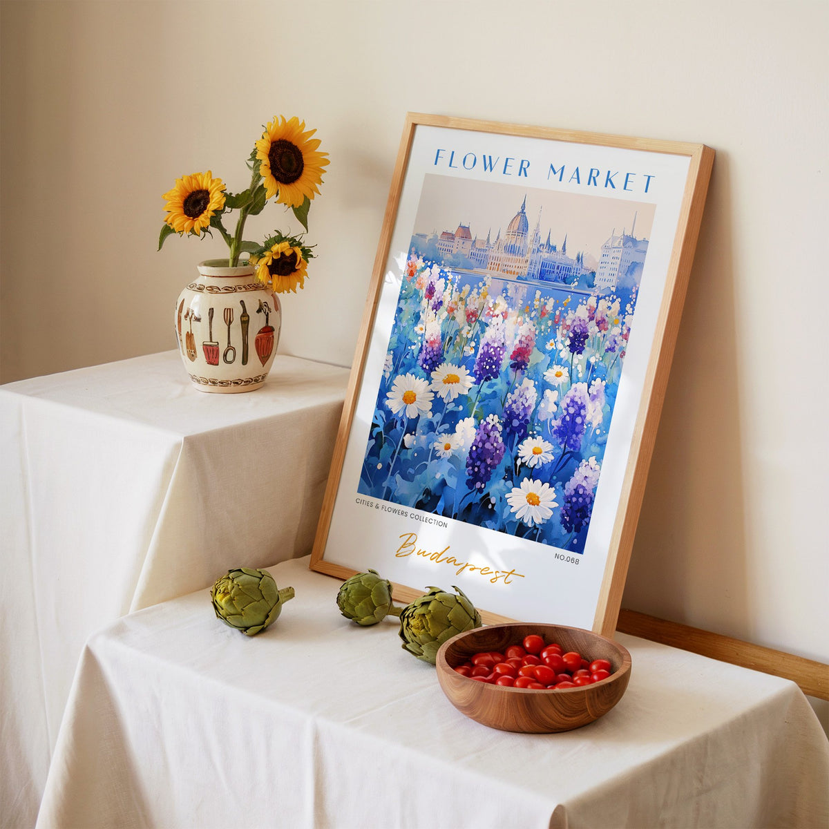Budapest flower market print in an oak frame leaning on a linen-draped table beside a vase of sunflowers