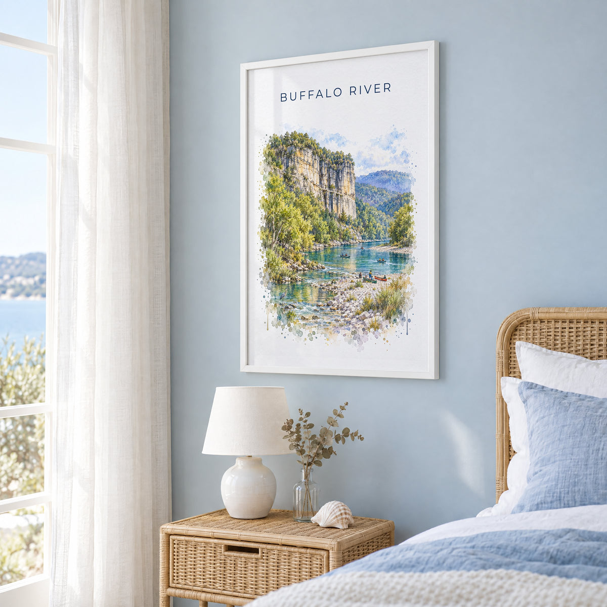 Buffalo National River watercolor print in a white frame on a pale blue wall beside a rattan bed in a coastal bedroom
