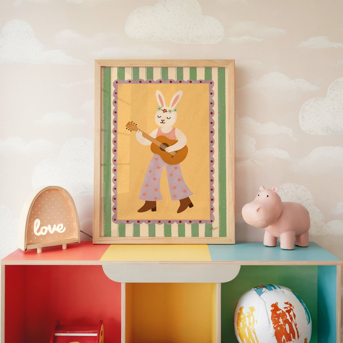 Bunny playing guitar with flower crown nursery print in an oak frame on a rainbow toy shelf against cloud wallpaper