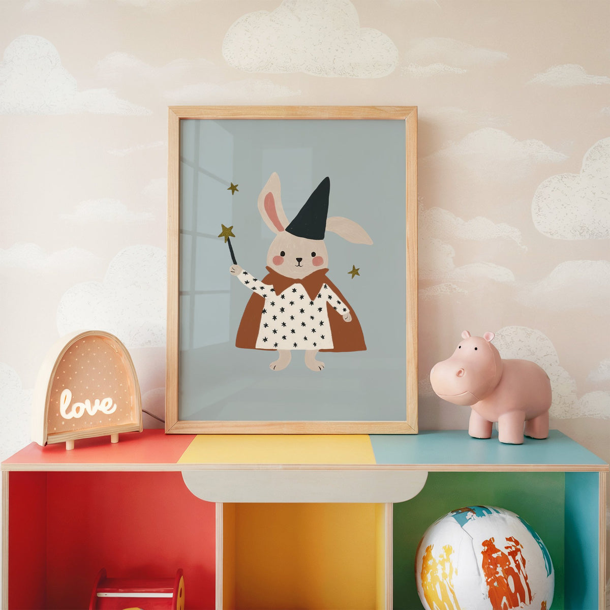 Bunny magician star wand nursery print in an oak frame on a rainbow toy shelf against cloud wallpaper
