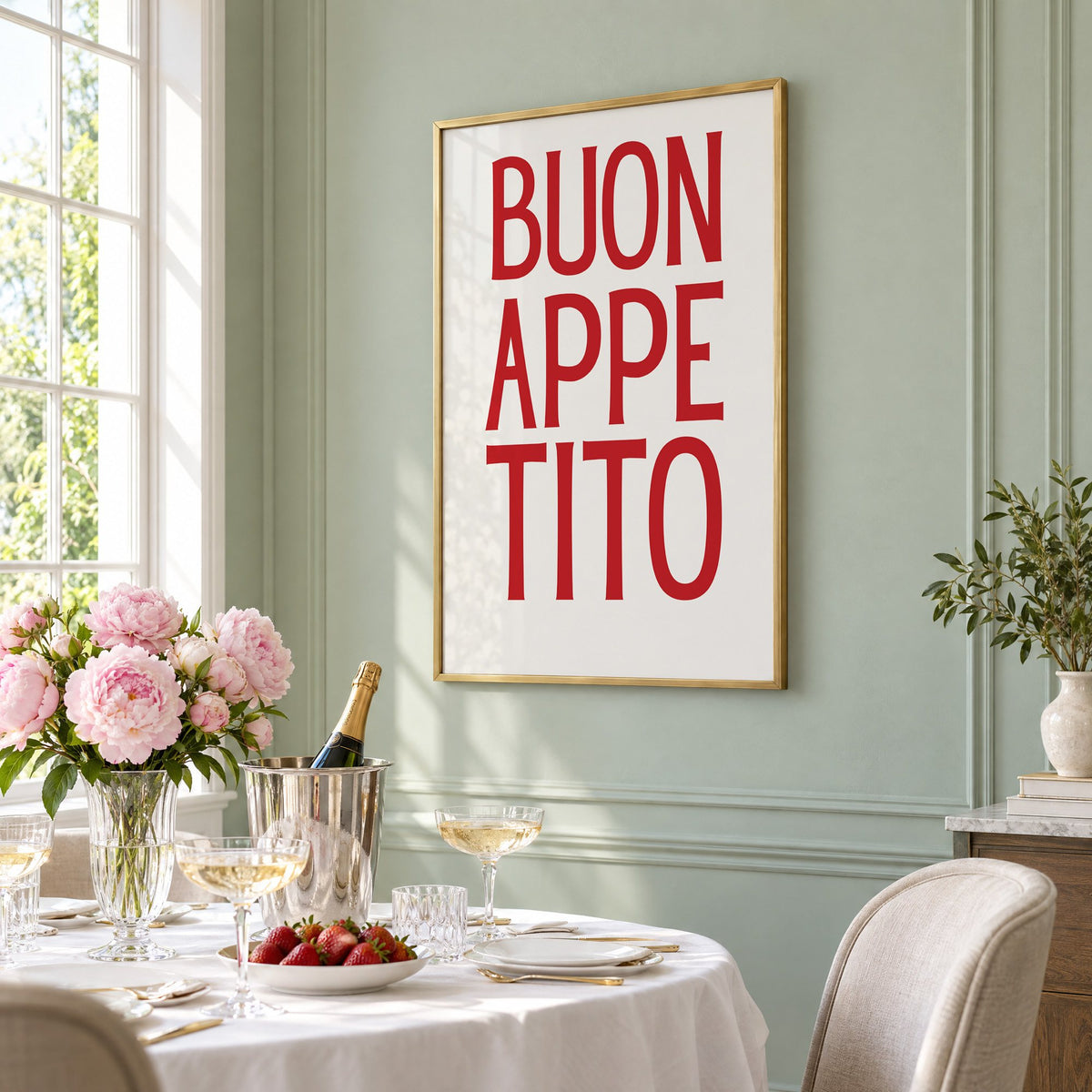 BUON APPE TITO red condensed type print in a brass frame on a sage wall above a brunch table with champagne and peonies, seen at an angle