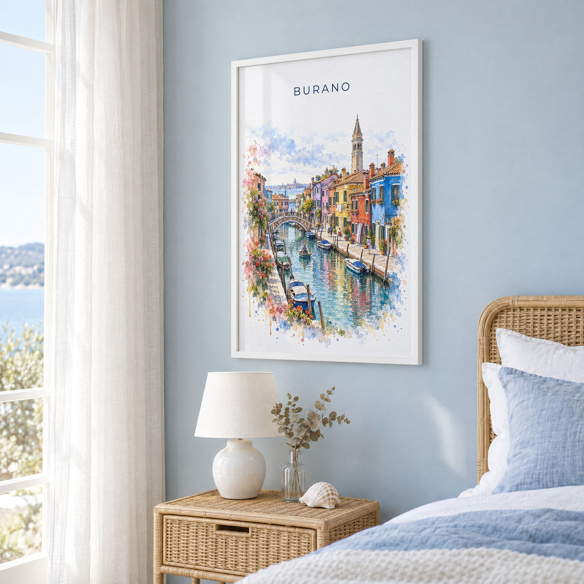 Burano watercolor print in a white frame on a pale blue wall beside a rattan bed in a coastal bedroom