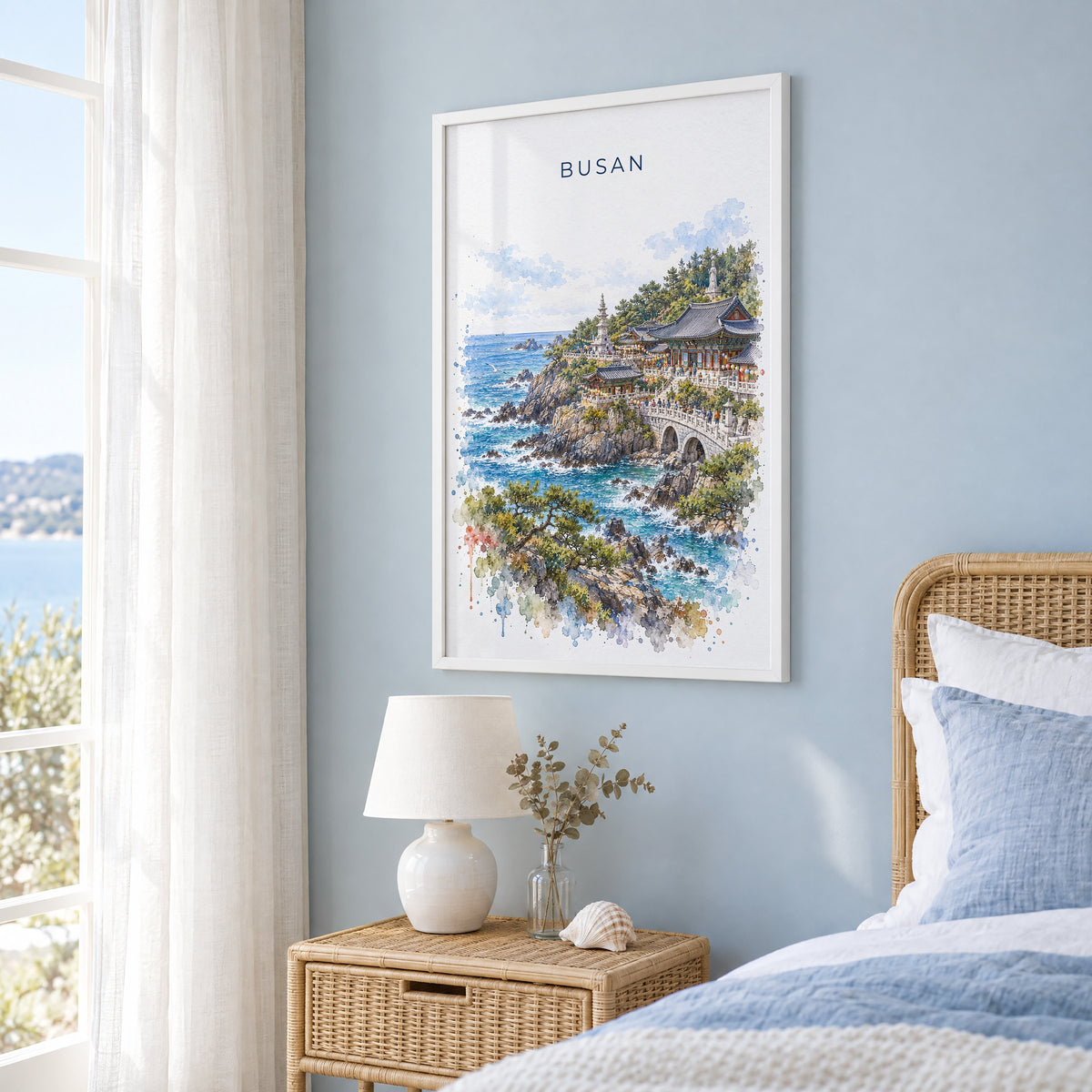 Busan watercolor print in a white frame on a pale blue wall beside a rattan bed in a coastal bedroom