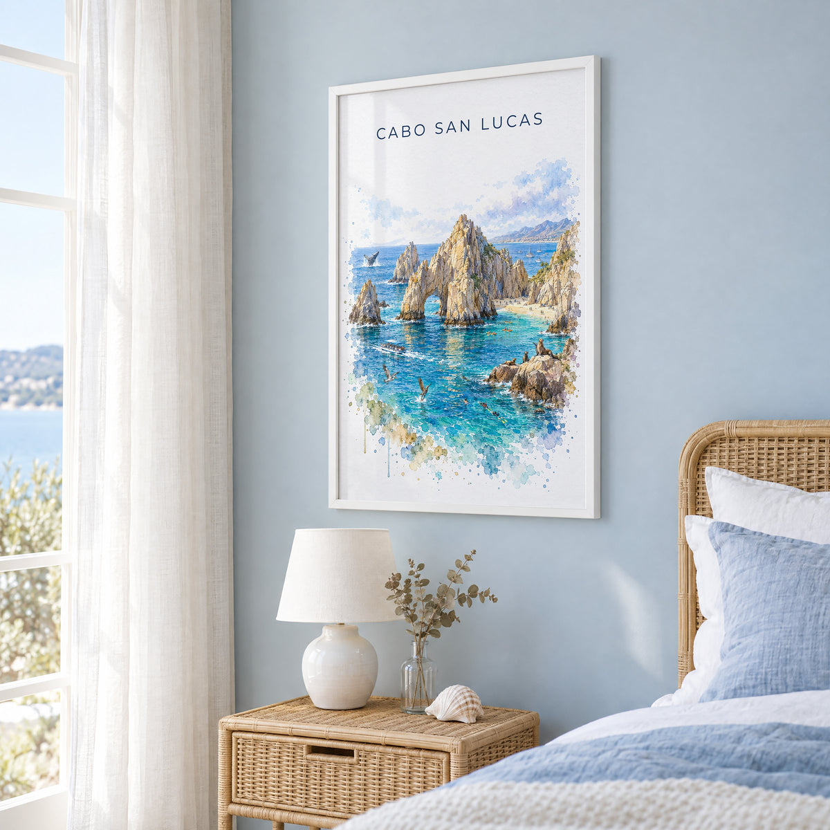 Cabo San Lucas watercolor print in a white frame on a pale blue wall beside a rattan bed in a coastal bedroom