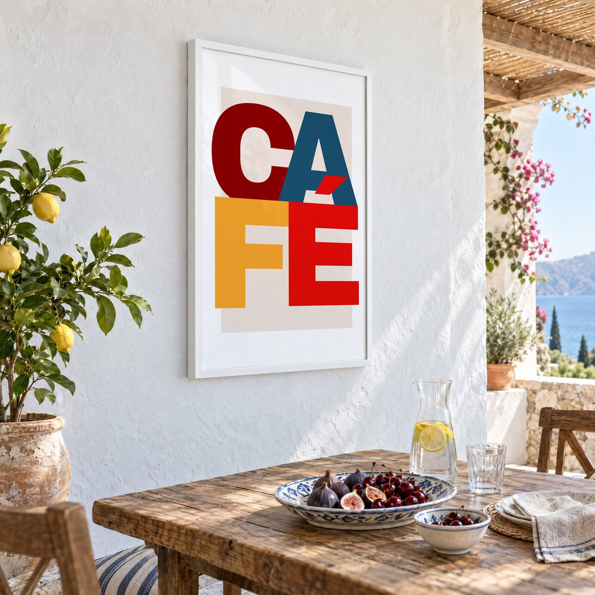 CA FÉ color block letters print in a white frame on a whitewashed terrace wall above a lunch table with figs and cherries, seen at an angle