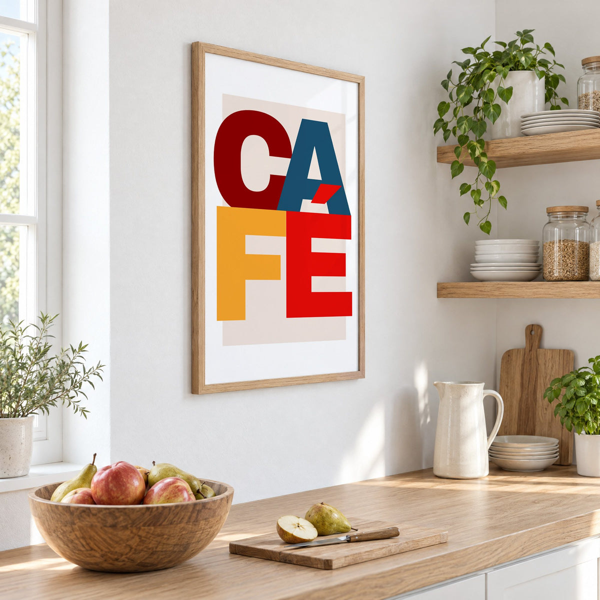 CA FÉ color block letters print in an oak frame on a white kitchen wall above a counter with apples and pears, seen at an angle