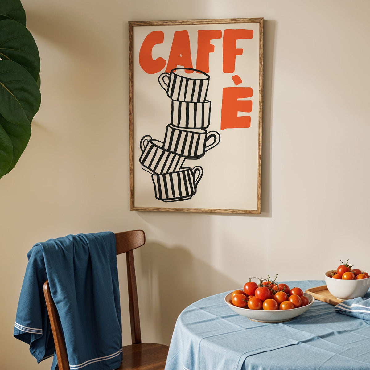 Stacked espresso cups print in an oak frame above a blue linen table with tomatoes