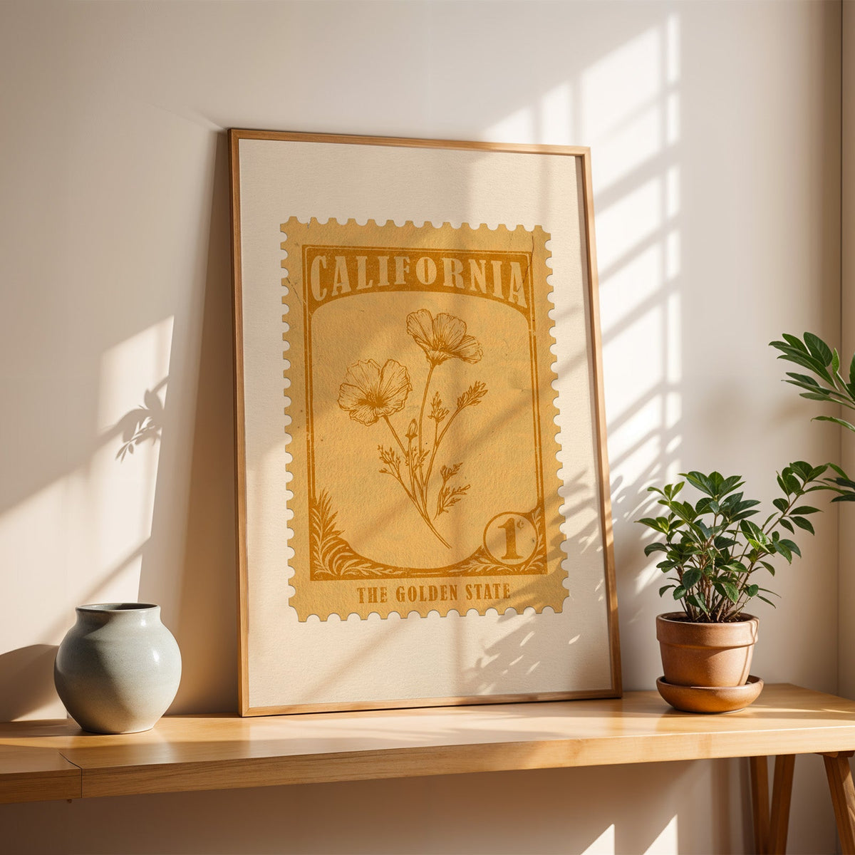 California poppy stamp print in a light oak frame leaning on a wooden table beside a stoneware vase