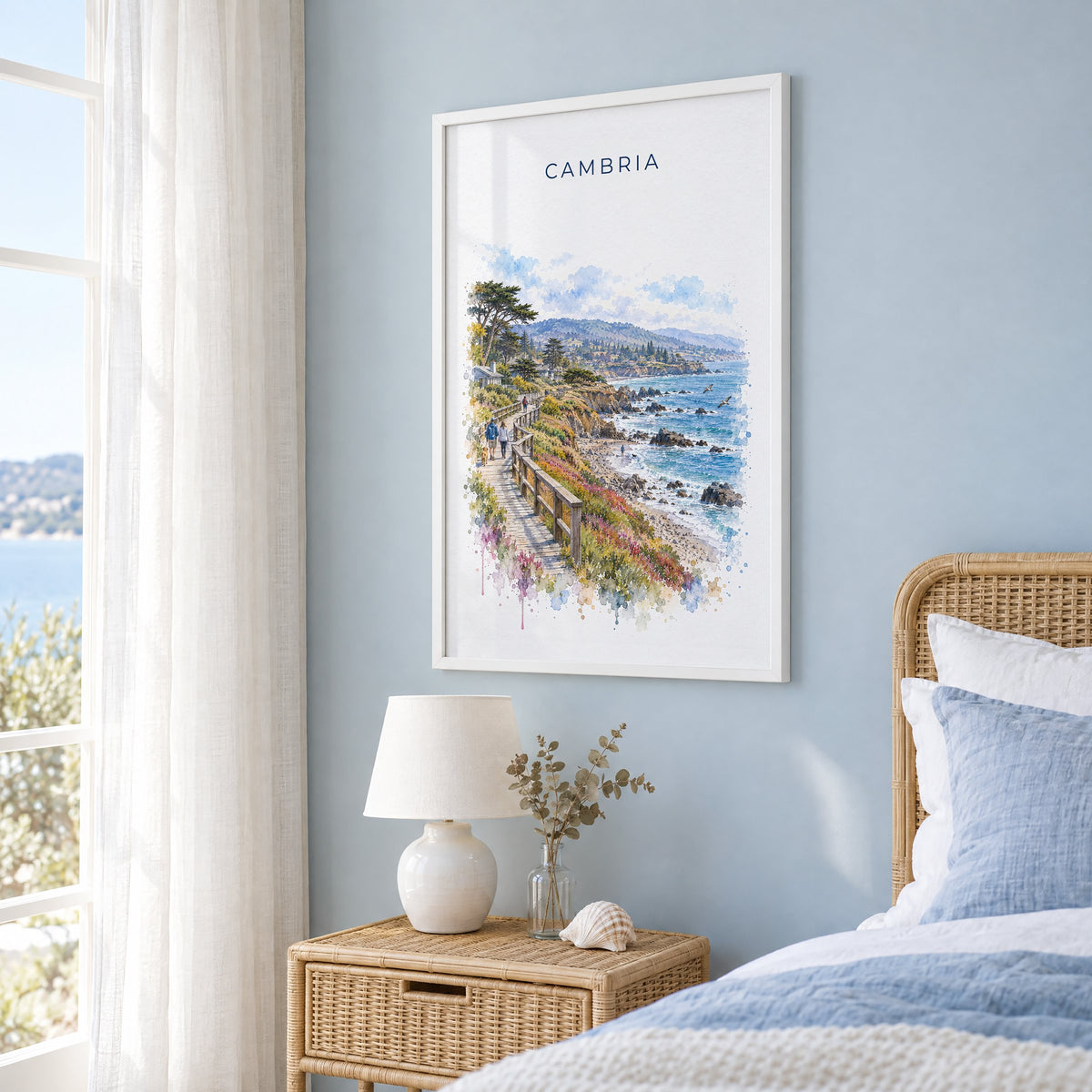 Cambria, California watercolor print in a white frame on a pale blue wall beside a rattan bed in a coastal bedroom