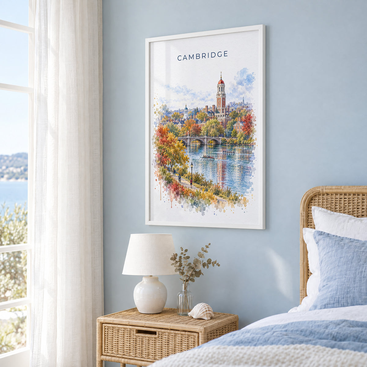 Cambridge, Massachusetts watercolor print in a white frame on a pale blue wall beside a rattan bed in a coastal bedroom