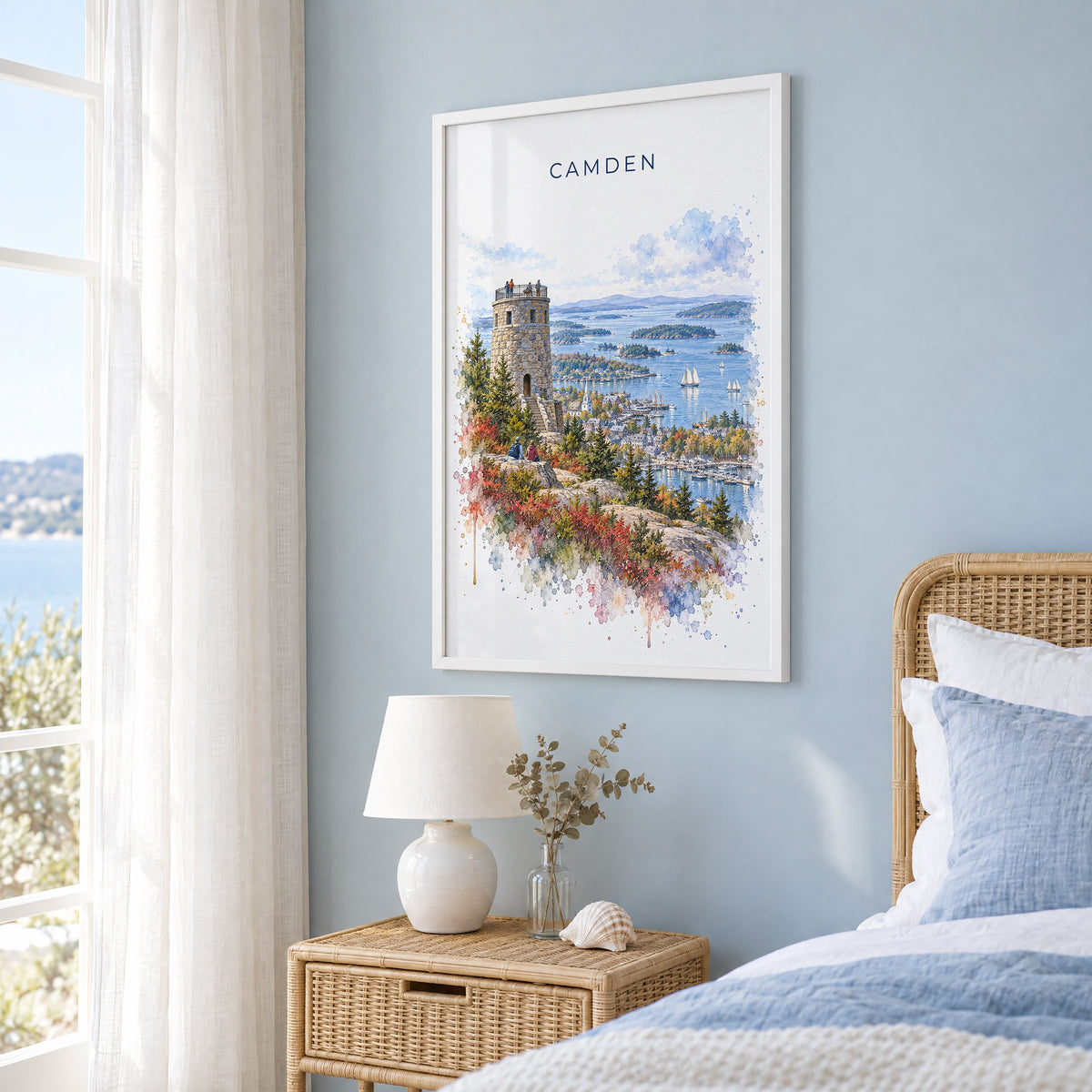 Camden, Maine watercolor print in a white frame on a pale blue wall beside a rattan bed in a coastal bedroom