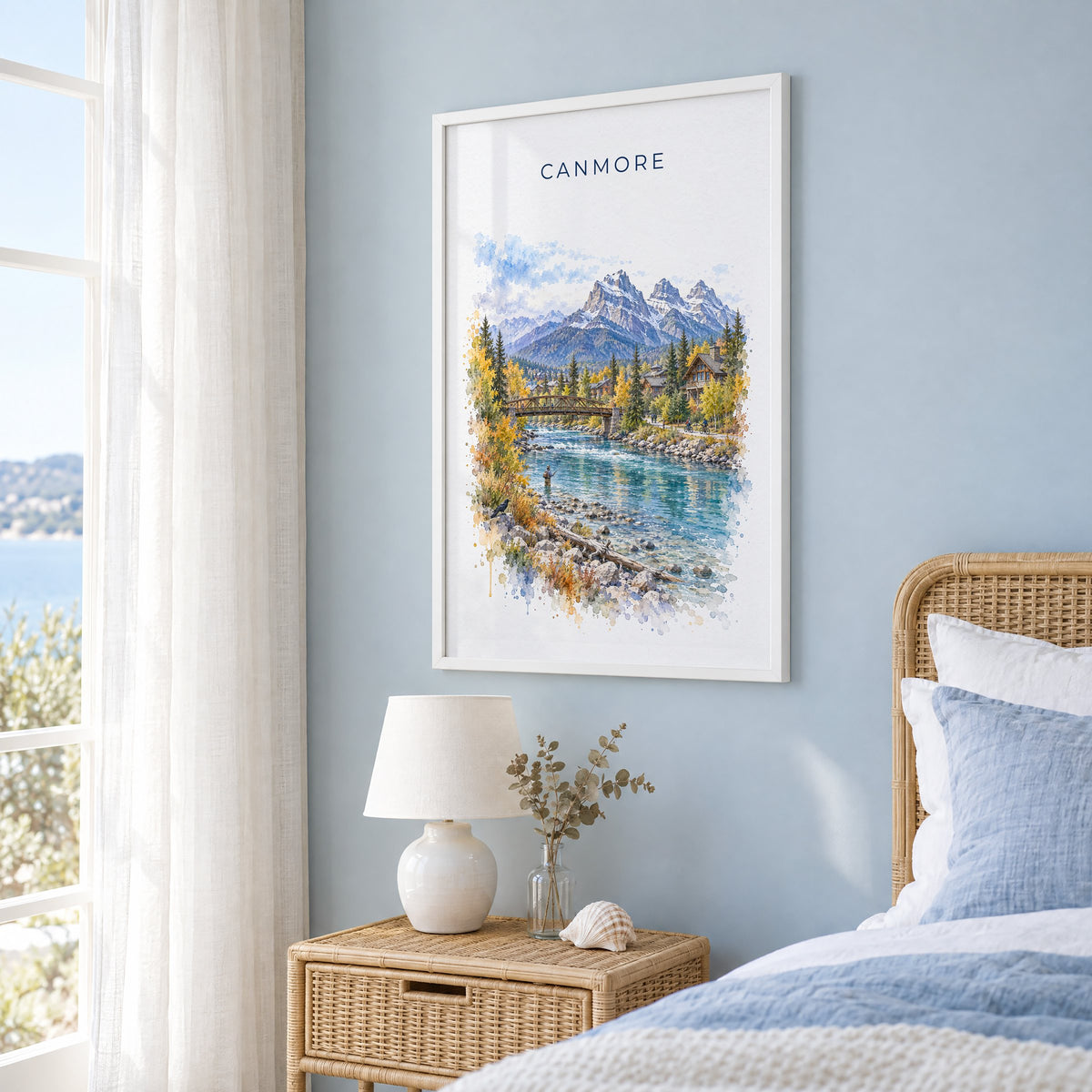 Canmore watercolor print in a white frame on a pale blue wall beside a rattan bed in a coastal bedroom