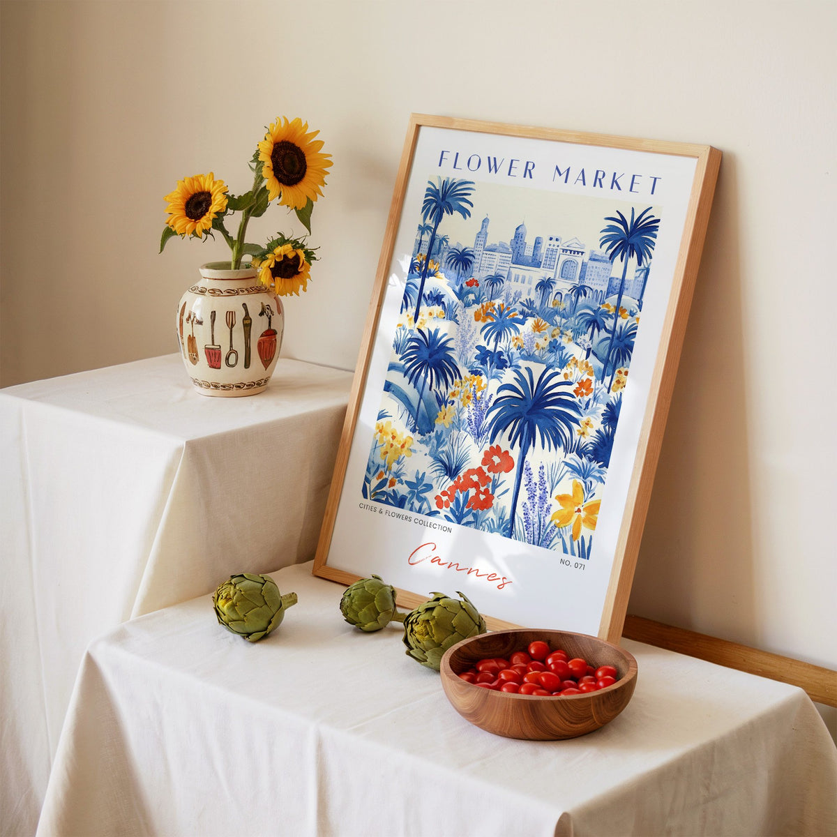 Cannes flower market print in an oak frame leaning on a linen-draped table beside a vase of sunflowers