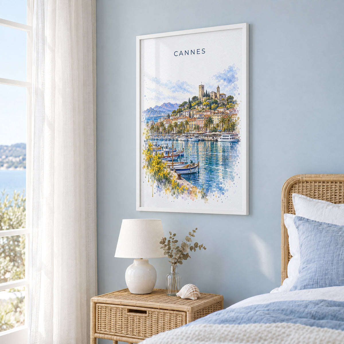 Cannes watercolor print in a white frame on a pale blue wall beside a rattan bed in a coastal bedroom