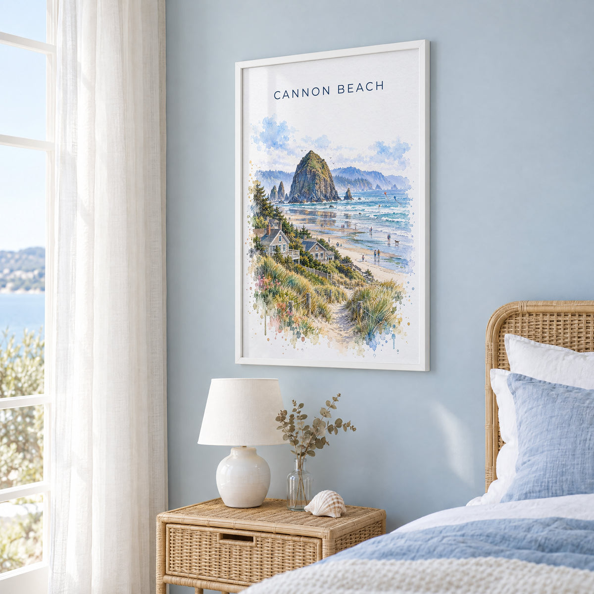Cannon Beach watercolor print in a white frame on a pale blue wall beside a rattan bed in a coastal bedroom