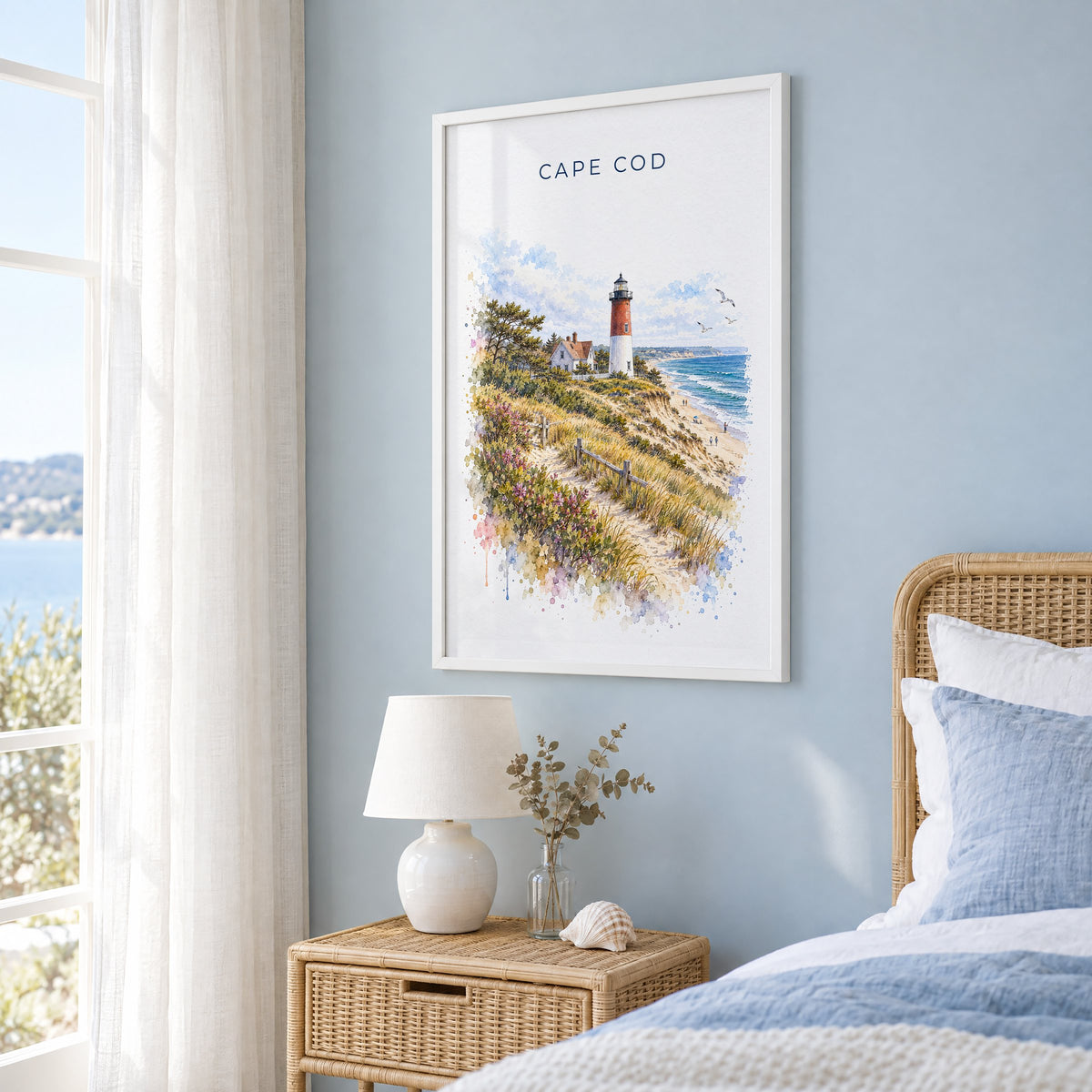 Cape Cod National Seashore watercolor print in a white frame on a pale blue wall beside a rattan bed in a coastal bedroom