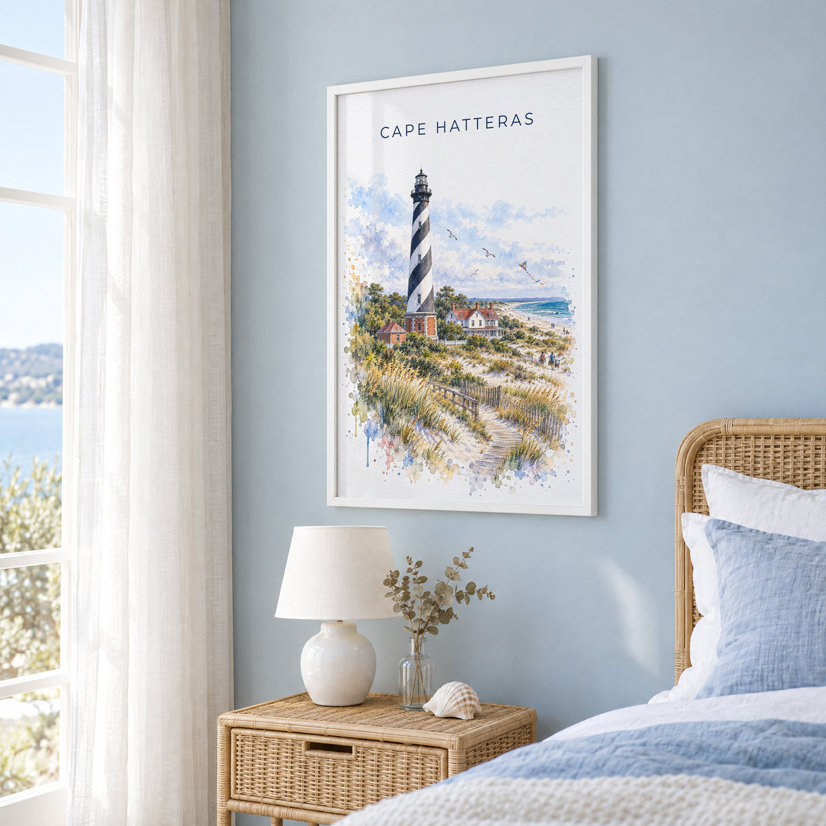 Cape Hatteras watercolor print in a white frame on a pale blue wall beside a rattan bed in a coastal bedroom